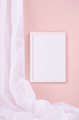 Delicate home decor with blank rectangle photo frame hanging on pastel pink wall, flow of silk curtain, vertical. Mock up for portfolio, design or text.