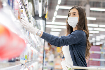Beautiful woman wearing medical face mask and rubber glove holding grocery basket picking up daily milk on product shelf. shopping at supermarket in new normal lifestyle concept during Coronavirus.