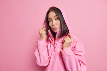 Serious self confident millennial girl with dark hair wears anorak hood on head looks directly at camera models against pink background going to have walk during cold windy day. People and style © WHstudio Leushin N