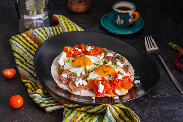 Huevos rancheros, Mexican fried egg on a wheat tortilla with tomato salsa, bean paste and feta cheese on a black plate on a light concrete background.