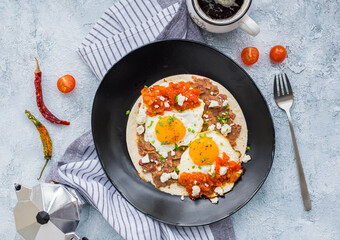 Huevos rancheros, Mexican fried egg on a wheat tortilla with tomato salsa, bean paste and feta cheese on a black plate on a light concrete background.