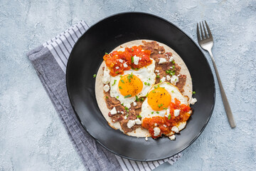 Huevos rancheros, Mexican fried egg on a wheat tortilla with tomato salsa, bean paste and feta cheese on a black plate on a light concrete background.