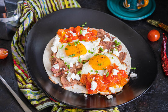 Huevos rancheros, Mexican fried egg on a wheat tortilla with tomato salsa, bean paste and feta cheese on a black plate on a light concrete background.