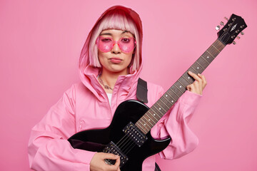 Stylish hipster woman wears trendy sunglasses jacket with hood poses with black electric guitar looks seriously at camera creats new song for her album isolated over pink background. Music concept