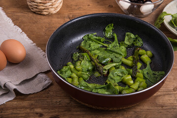 Cooking omelet with asparagus, tomatoes and spinach - fried spinach and asparagus in a frying pan on a wooden table, eggs and tomatoes with cheese in a cup