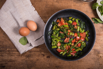Cooking omelet with asparagus, tomatoes and spinach - fried asparagus, tomatoes and spinach in a frying pan on a wooden table and eggs