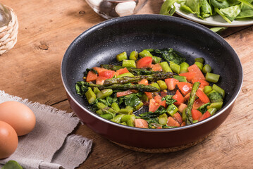 Cooking omelet with asparagus, tomatoes and spinach - fried asparagus, tomatoes and spinach in a frying pan on a wooden table and eggs
