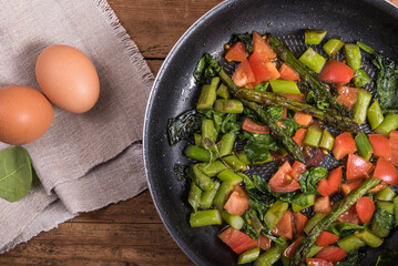 Cooking omelet with asparagus, tomatoes and spinach - fried asparagus, tomatoes and spinach in a frying pan on a wooden table and eggs