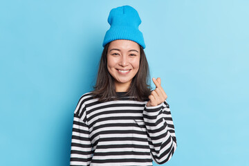 Waist up shot of pretty Asian woman with dark hair makes korean heart with fingers wears hat striped jumper smiles pleasantly isolated over blue background expresses love. Body language concept