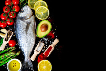 Raw sea bream fish arranged on black background with fresh vegetables and spices