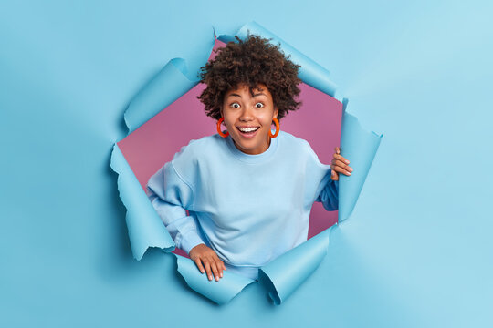 Happy Surprised Afro American Teenage Girl Reacts On Something Awesome Smiles Broadly Poses In Torn Paper Blue Wall Dressed In Casual Jumper Focused At Camera. Human Reactions And Emotions Concept