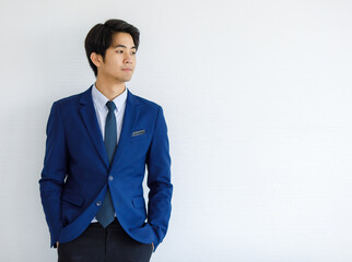 Portrait closeup shot of Asian young handsome happy intelligence college teen student wears blue formal suit and necktie standing hold hands in pants pockets look outside in front of white wall