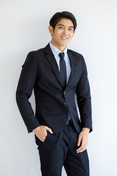 Portrait closeup shot of Asian young handsome happy management lawyer officer in formal black suit and necktie standing hold hands in pants pockets against white wall background