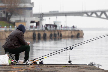 A fisherman with a phone in his hands and a hood in the spring sits on the Volga River in Russia Saratov against the background of a bridge on the embankment of cosmonauts and looks into a smartphone.