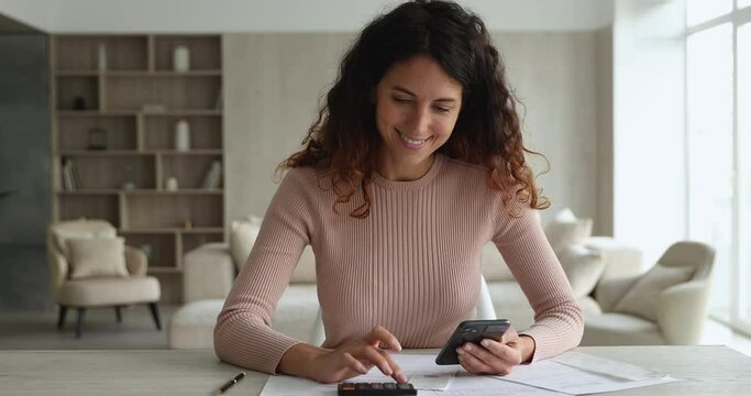 Woman sit at desk makes calculations on calculator writes results, use e-bank app on cell, pay bills through online bank. E-pay, personal finances management, lead business, calculating income concept