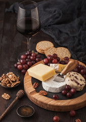 Glass of red wine with selection of various cheese on the round board and grapes on wooden background. Blue Stilton, Red Leicester and Brie Cheese and bowl of nuts and crackers. Top view