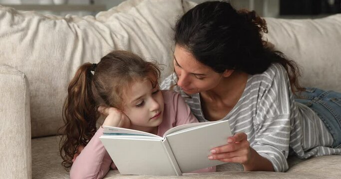 Close up caring young mum her preschool pretty daughter lying on comfy sofa at home with fairytale, mum read book to kid girl. Offspring development, stories for children to teach life values concept