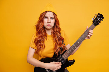 Dissatisfied offended redhead European woman purses lower lip plays electric guitar dressed casually poses with musical instrument isolated over yellow background. Music and singing concept.