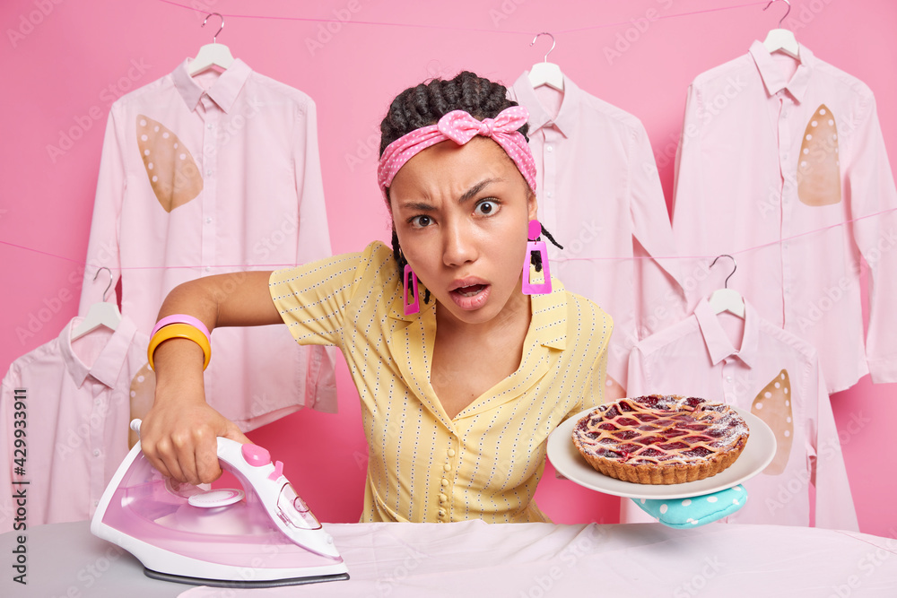 Wall mural Serious attentive young housewife concentrated directly at camera uses electric irone for stroking laundry busy cooking delicious pie for family wears headband and dress engaged in domestic work