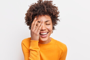 Fototapeta na wymiar Close up shot of joyful carefree young woman with curly Afro hair smiles toothily keeps eyes closed makes face palm wears orange jumper expresses happiness isolated over white studio background