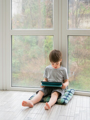 Curious boy watch cartoons on digital tablet. Kid sits on floor and uses electronic device. Indoor leisure for children while it's raining outside.