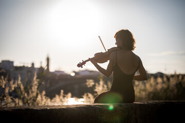 Young woman violinist posing playing on the street during sunset against light