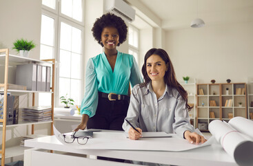 Two happy beautiful stylish women at work. Indoor portrait of black and caucasian coworkers, professional designers or architects, looking at camera. Successful career and diverse workplace concept