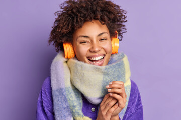 Close up shot of happy young African American woman keeps hands together smiles broadly wears scarf around neck uses wireless headphones for listening music enjoys good sound isolated on purple