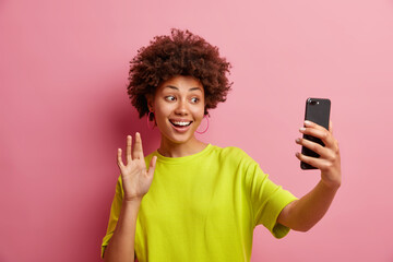 Cheerful young Afro American woman with cury hair waves in smartphone camera makes hi gesture while...