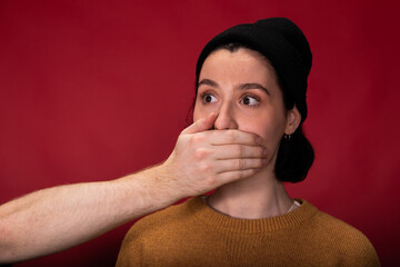 Speak no, mute, freedom of speech and voice, silence, subordination, me too concept. Young hipster woman standing with closed mouth by male hand on isolated red background. Dependent female person