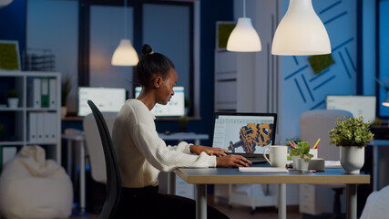 African engineer working late at night on a 3D model of industrial gears at laptop doing overtime...