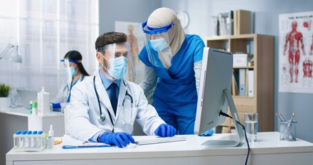 Portrait of mixed-race hospital workers working in cabinet. Caucasian male doctor typing on computer sitting at desk working with Arab young female nurse. Healthcare, coronavirus pandemic concept