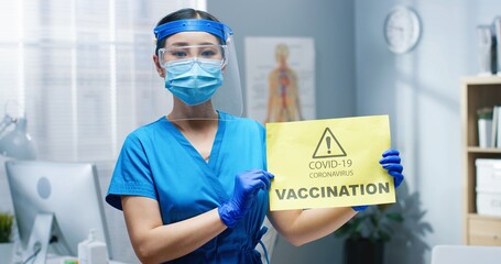 Close up portrait of Asian young healthcare worker nurse in protective face shield and mask holding in hands in gloves card saying Covid-19 vaccination. notification about coronavirus vaccine