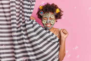 Positive young Afro American woman applies beauty mask with cucumber slices clenches fist has fun while taking shower hides behind curtain poses against pink background has clean refeshed body