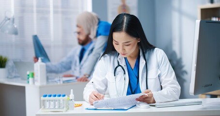 Portrait of Asian beautiful young female professional doctor in white medical coat sitting at desk...