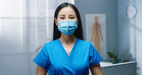 Asian young beautiful female professional nurse wearing medical mask and uniform standing in hospital at work looking at camera in good mood working during coronavirus pandemic, clinic, close up