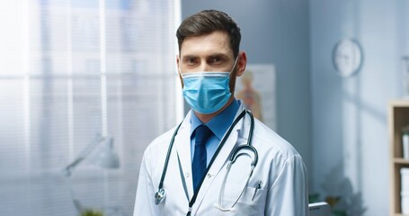Close up of happy Caucasian young handsome man general practitioner in medical mask standing in medical center looking at camera and smiling. Male doctor at work during covid-19 pandemic
