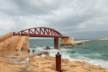 Beautiful Malta sea shore pier view with red bridge on rainy cloudy day with big white waves , yellow sandstone rocks, shrubs, blue water, and skies. white fence, water