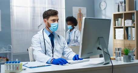Fototapeta na wymiar Portrait of Caucasian young handsome man professional physician at workplace in coronavirus pandemic sitting at table in cabinet texting on computer browsing online looking at monitor Hospital concept