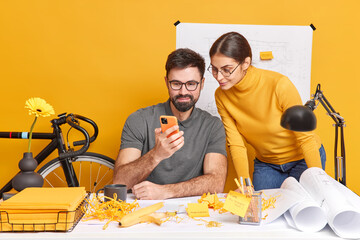 Professional architects look attentively at smartphone try to improve productivity of work pose at coworking space with papers blueprints share creative ideas with each other. Designers at workplace