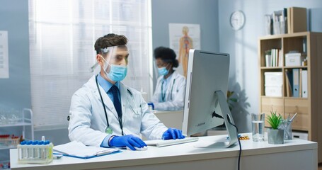 Portrait young Caucasian male doctor sitting in cabinet in medical center during covid quarantine typing on computer and reading from screen Man physician searching internet at work Hospital concept
