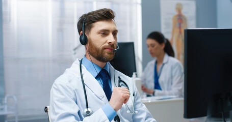 Close up portrait of Caucasian young handsome male physician specialist speaking on video call in headset on computer sitting in hospital cabinet showing vaccine cure. Medical video consultation