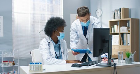 Obraz premium Mixed-race male and female doctors in medical masks speaking discussing analysis results working in hospital. African American young woman physician talking to Caucasian coworker. Healthcare concept