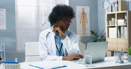 African American young pretty woman professional physician in white medical coat tapping on laptop computer searching internet looking at monitor sitting at desk in cabinet, hospital, Portrait concept