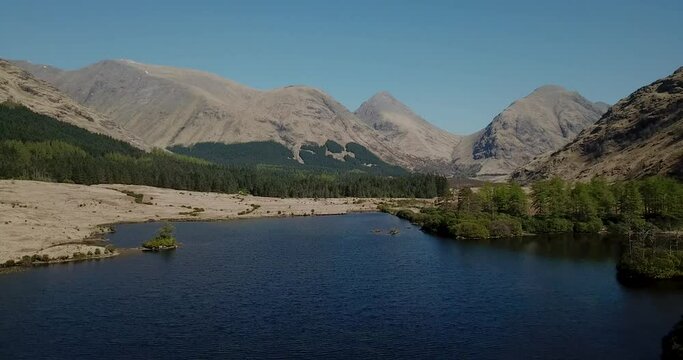 4k aerial footage of a tranquil loch in Glen Etive, Scottish Highlands with mountains and cloudless blue sky backdrop. Small island of trees in loch. No people.