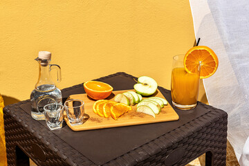 Close up photo of the small brown sunlit table with glass of fresh orange  juice and carafe with white traditional cretan spirit raki and cut fruits: apple and orange. 