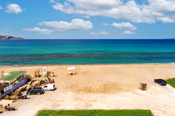 Seaside, with blue and green sea and light blue sky with white clouds, sandy beach and some cars parked nearby.