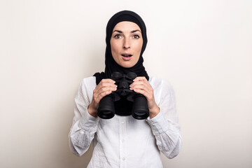 Friendly surprised Young Muslim woman in white shirt and hijab holds binoculars. Banner