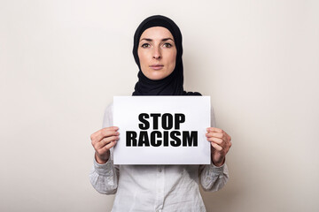 Muslim young woman in hijab holds a banner with the text STOP RACISM on a light background