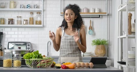 POV of a young, happy and beautiful Asian woman with curly long hair wearing grey gingham apron. She's makes video about making meal or live streaming online to show cooking method in modern kitchen.